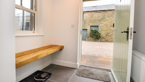 A clean, modern entryway with a wooden bench, a small shoe rack, and a door leading to a courtyard with stone walls and a potted plant. - 1 Hull House Cottage (Skipton)