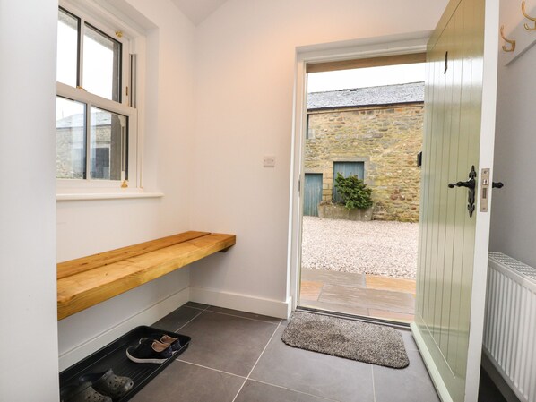 A clean, modern entryway with a wooden bench, a small shoe rack, and a door leading to a courtyard with stone walls and a potted plant. - 1 Hull House Cottage (Skipton)
