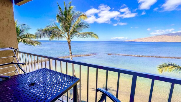 Un balcón con una mesa y sillas de metal, con vista a una playa con palmeras y un cielo azul despejado.