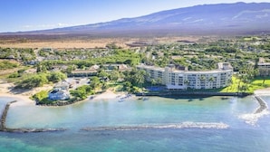 Vista aérea de un resort con un edificio grande rodeado por una playa, agua cristalina y un paisaje montañoso.