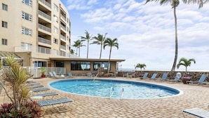 Un área de piscina con sillones de descanso, palmeras y un edificio con balcones.