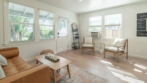 Una sala de estar con un sofá, una mesa de centro y dos sillas. Hay ventanas grandes con persianas, una escalera y un estante montado en la pared.
