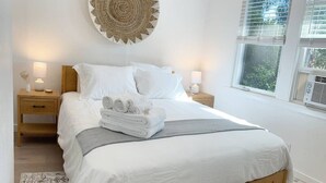 Una cama bien tendida con sábanas blancas y una manta gris, dos mesitas de noche con lámparas, una pieza decorativa tejida en la pared y una ventana con persianas.