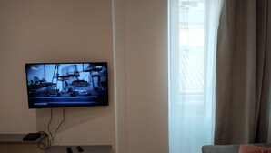 A living room with a flat-screen TV mounted on the wall, a sofa, and a window with sheer curtains. - Charming studio downtown Ajaccio (Ajaccio)