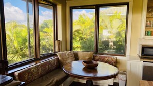 A corner seating area with a round table and a bowl, large windows with a tropical view, and a microwave on the counter. - Walk to Poipu Beach • 4BR + Bonus • Pool (Koloa)