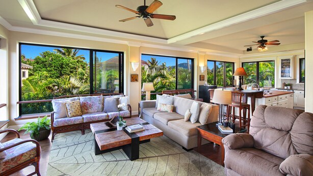 A spacious living area with a sofa set, a coffee table, and a dining table. There are ceiling fans and large windows offering a view of greenery outside. - Walk to Poipu Beach • 4BR + Bonus • Pool (Koloa)