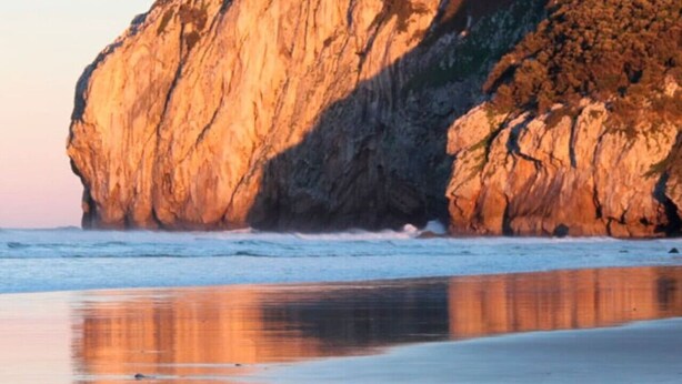 Una gran formación rocosa junto al mar, con una playa arenosa al frente.