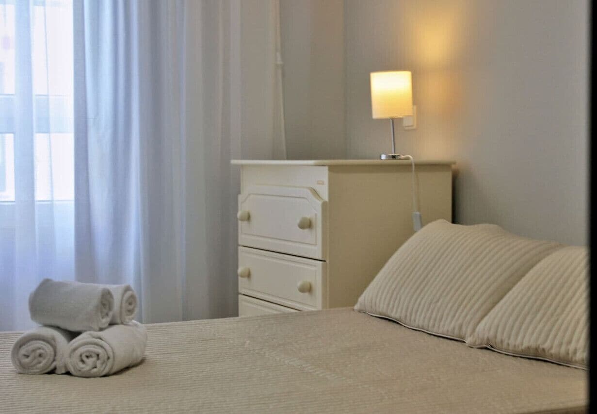 A neatly made bed with rolled white towels, a bedside lamp, and a white dresser in a hotel room.