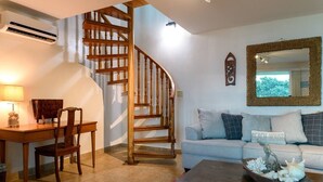 A living room with a wooden staircase, a sofa with multiple cushions, a desk with a chair, and a decorative bowl on a coffee table. - Merizo Seaside B&B - Unit 2 Coral Suite (Tasi Merizo)