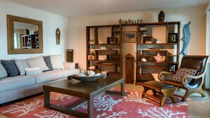 A living room with a sofa, coffee table, and a chair. There is a bookshelf with various items and a mirror on the wall. - Merizo Seaside B&B - Unit 2 Coral Suite (Tasi Merizo)