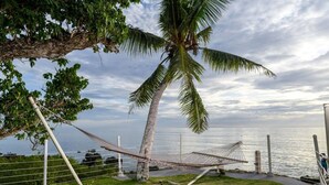 A hammock set up on a grassy area with a view of the sea, a palm tree, and a fence. - Merizo Seaside B&B - Unit 3 Ocean Villa (Tasi Merizo)