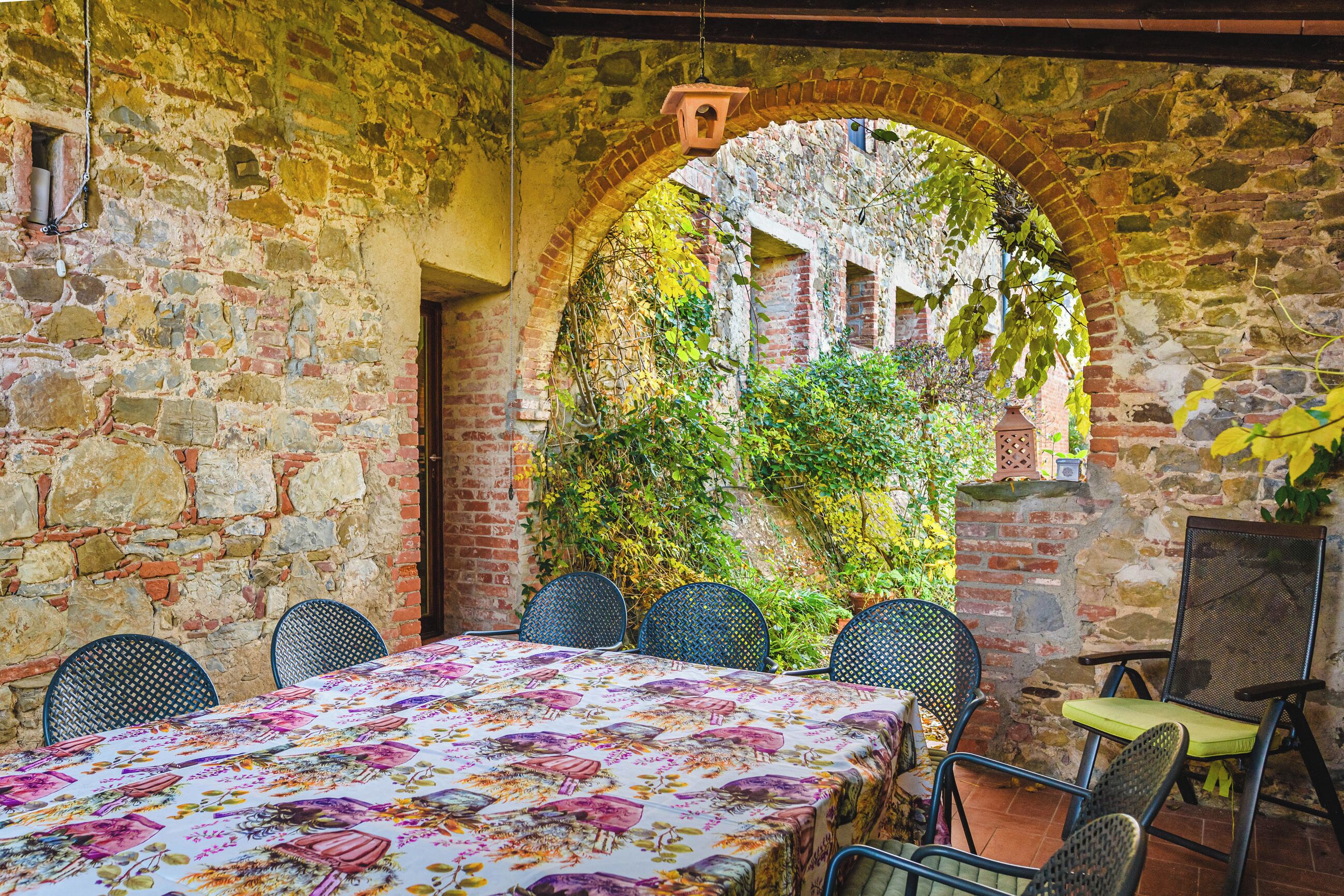 A rustic stone-walled room with a table set for six, surrounded by chairs, and an arched doorway leading to a garden area.