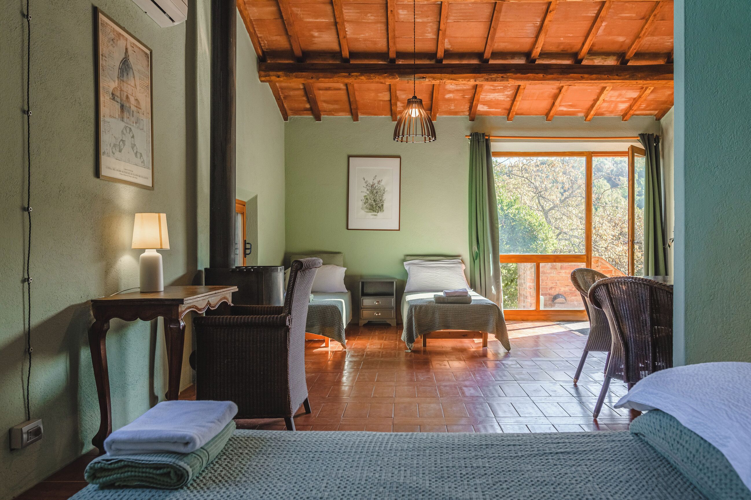 A cozy room with two beds, a wooden ceiling, a window with a view, and a lamp on a table.
