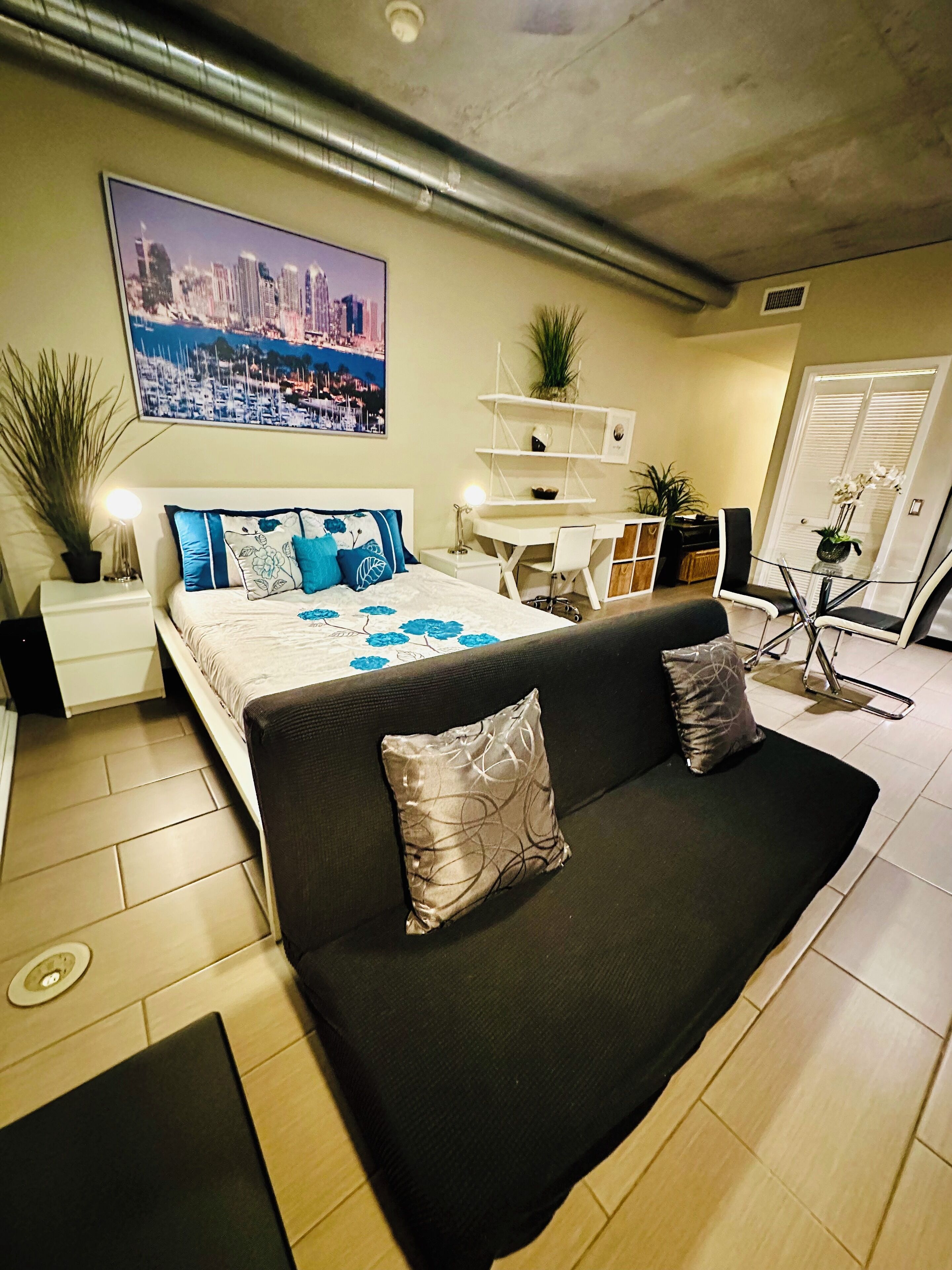A modern bedroom with a bed, a black sofa, a desk, and a cityscape painting on the wall.