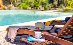 Una silla de madera para relajarse con un sombrero, un libro y una taza con lunares sobre una mesita junto a una piscina.