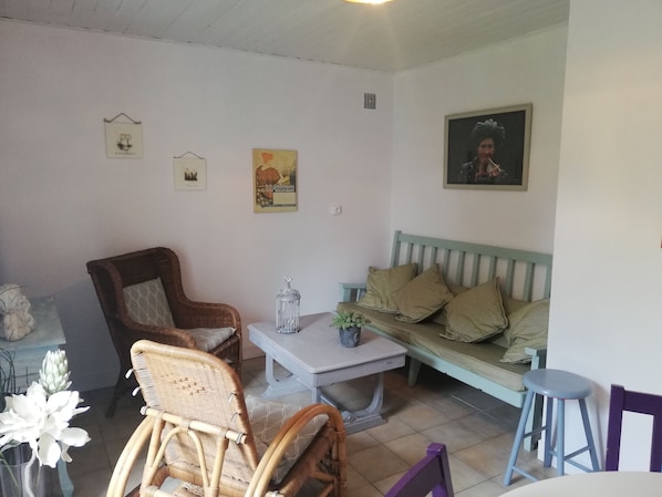Ein gemütliches Wohnzimmer mit einem braunen Korbstuhl, einem hellblauen Sofa mit gemusterten Kissen, einem kleinen Tisch mit einem Glas und einem gerahmten Bild an der Wand.