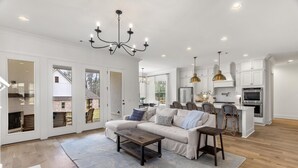 Amplia sala de estar con un sofá blanco grande, una mesa de centro de madera y un candelabro. Incluye un área de cocina con armarios blancos y un comedor con mesa y sillas.