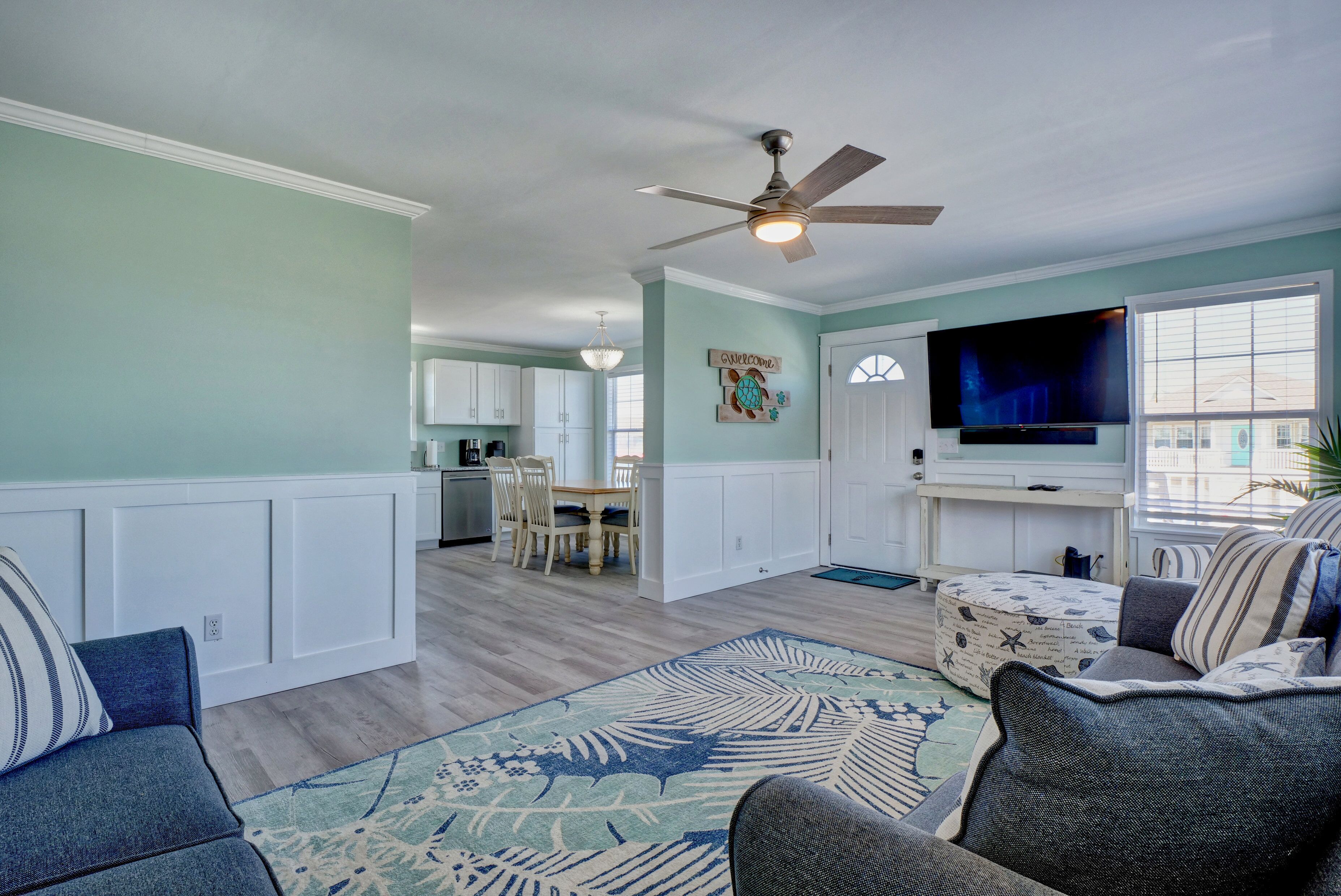 A spacious living room with a large TV, comfortable seating, a ceiling fan, and a view of the kitchen.