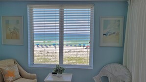 A room with a large window overlooking a beach, a chair, a small table with a plant, and two framed pictures on the wall.