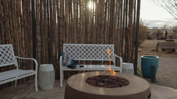 Un espace extérieur aménagé avec un foyer, des bancs en métal blanc et une clôture en bambou tressé.