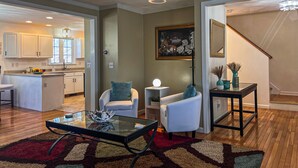 A spacious living area with a glass coffee table, two white armchairs, a small side table with a lamp, and a painting on the wall.