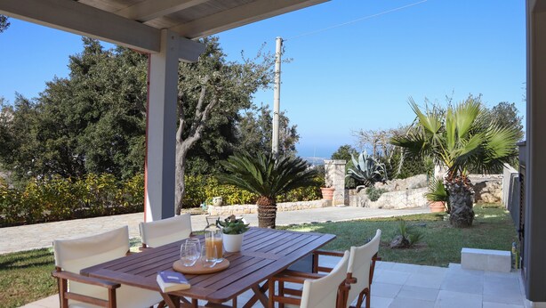 A covered outdoor patio with a wooden table and chairs, surrounded by greenery and a view of the sea.