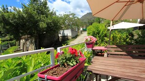 Una terraza de madera con un banco y una mesa, rodeada de plantas en macetas y un camino que lleva a un edificio.