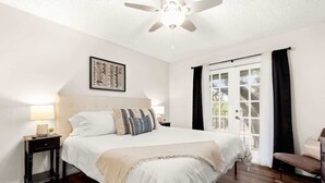A bedroom with a bed, a nightstand, a ceiling fan, a window with curtains, and a door. - Heated Pool-HotTub-Game Room-Fire Pit-Big Backyard (San Antonio)