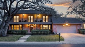 A two-story house with a garage, a well-maintained lawn, and a large tree in the foreground. - Heated Pool-HotTub-Game Room-Fire Pit-Big Backyard (San Antonio)