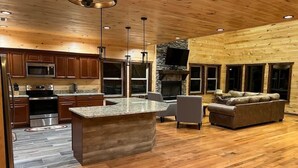 A spacious living area with a fireplace, wooden flooring, and a kitchen with granite countertops and modern appliances. - Luxury Mtn View Cabin w/ Theater, Hot Tub, Game Room, EV Charger, Sleeps 12 (Gatlinburg)