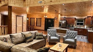 A spacious living area with a leather sofa, two armchairs, and a coffee table. The room features wooden walls and ceiling, a kitchen with modern appliances, and a dining area with a table and chairs. - Luxury Mtn View Cabin w/ Theater, Hot Tub, Game Room, EV Charger, Sleeps 12 (Gatlinburg)