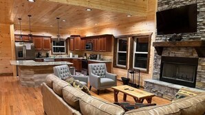 A spacious living area with a large leather sofa, two armchairs, a coffee table, and a fireplace. The room features wooden flooring, a stone accent wall, and a kitchen area with wooden cabinets and a stainless steel refrigerator. - Luxury Mtn View Cabin w/ Theater, Hot Tub, Game Room, EV Charger, Sleeps 12 (Gatlinburg)
