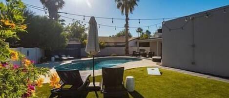 A backyard with a swimming pool, a lawn, and a patio area with chairs and an umbrella.