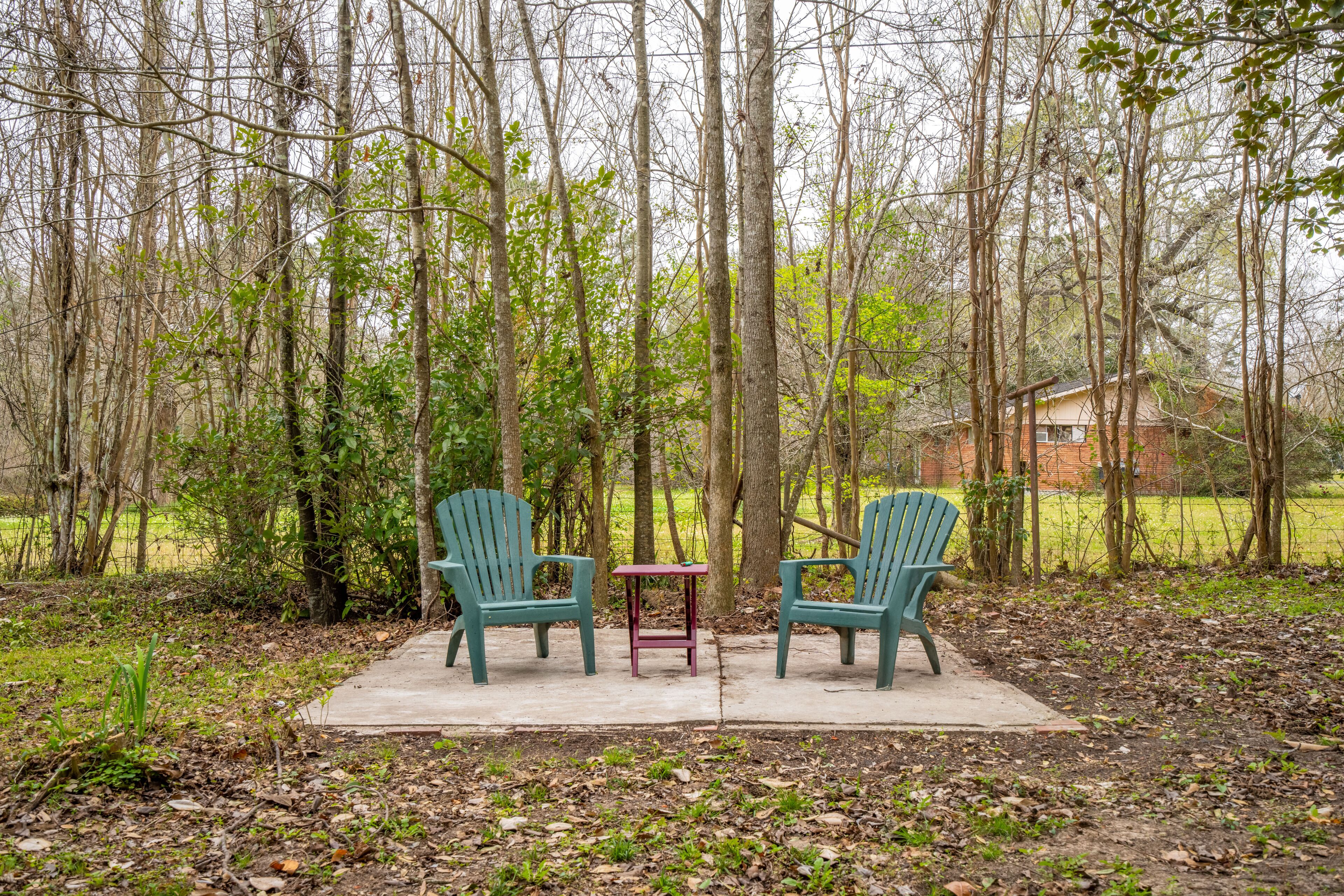 Two green Adirondack chairs and a small table in a forested area with a house in the background.