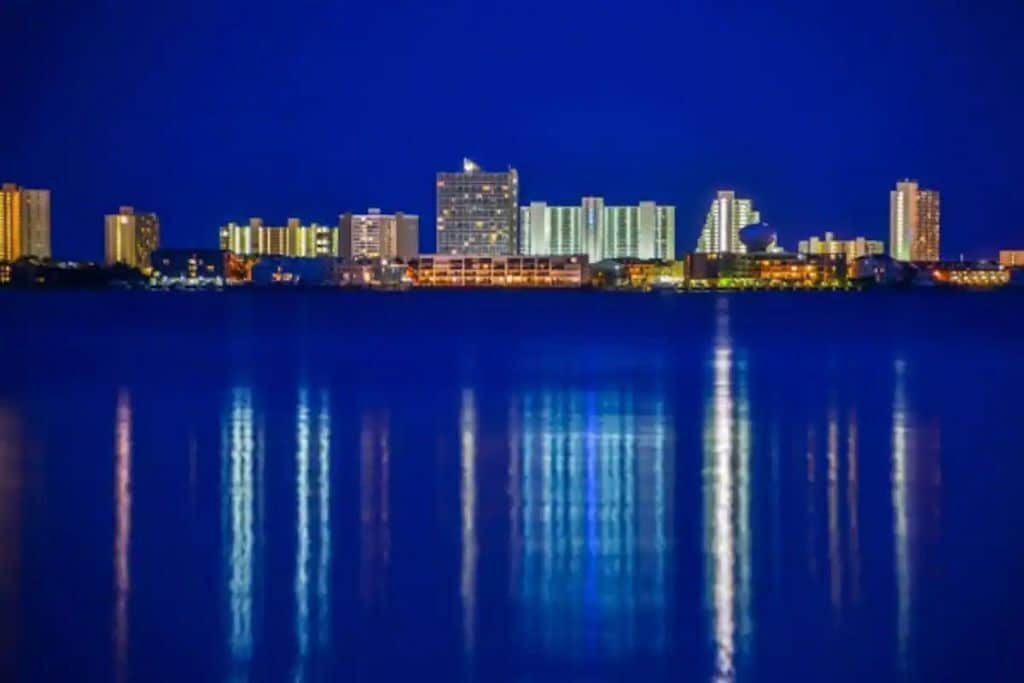 Une vue nocturne sur les grat-ciel d’une ville, les bâtiments illuminés se reflétant dans l’eau calme.