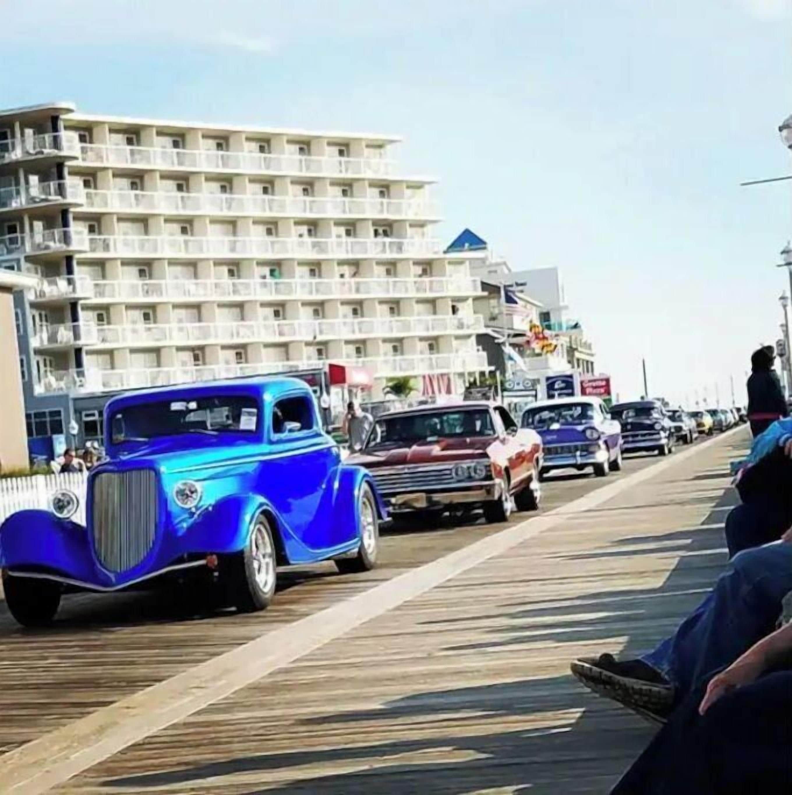 Ein blauer Oldtimer fährt auf einem Promenadenweg entlang, im Hintergrund ein mehrstöckiges Gebäude.