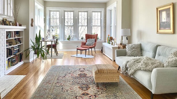 A spacious living room with hardwood floors, a large window, a red armchair, a grey sofa, and a bookshelf.
