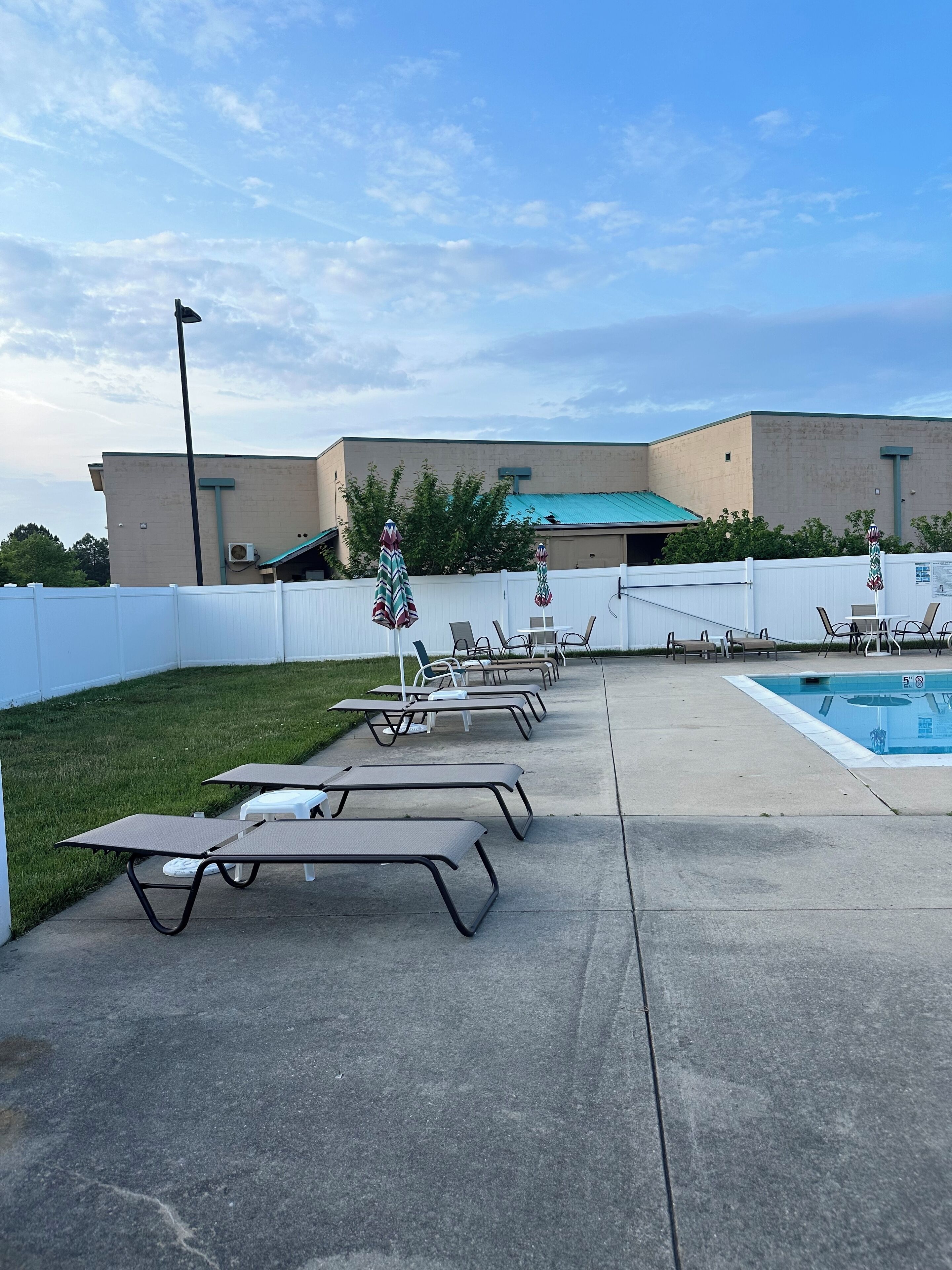 Un espace aménagé au bord de la piscine, avec des chaises longues et des parasols, une allée en béton et, au fond, un bâtiment doté d’un toit végétalisé.