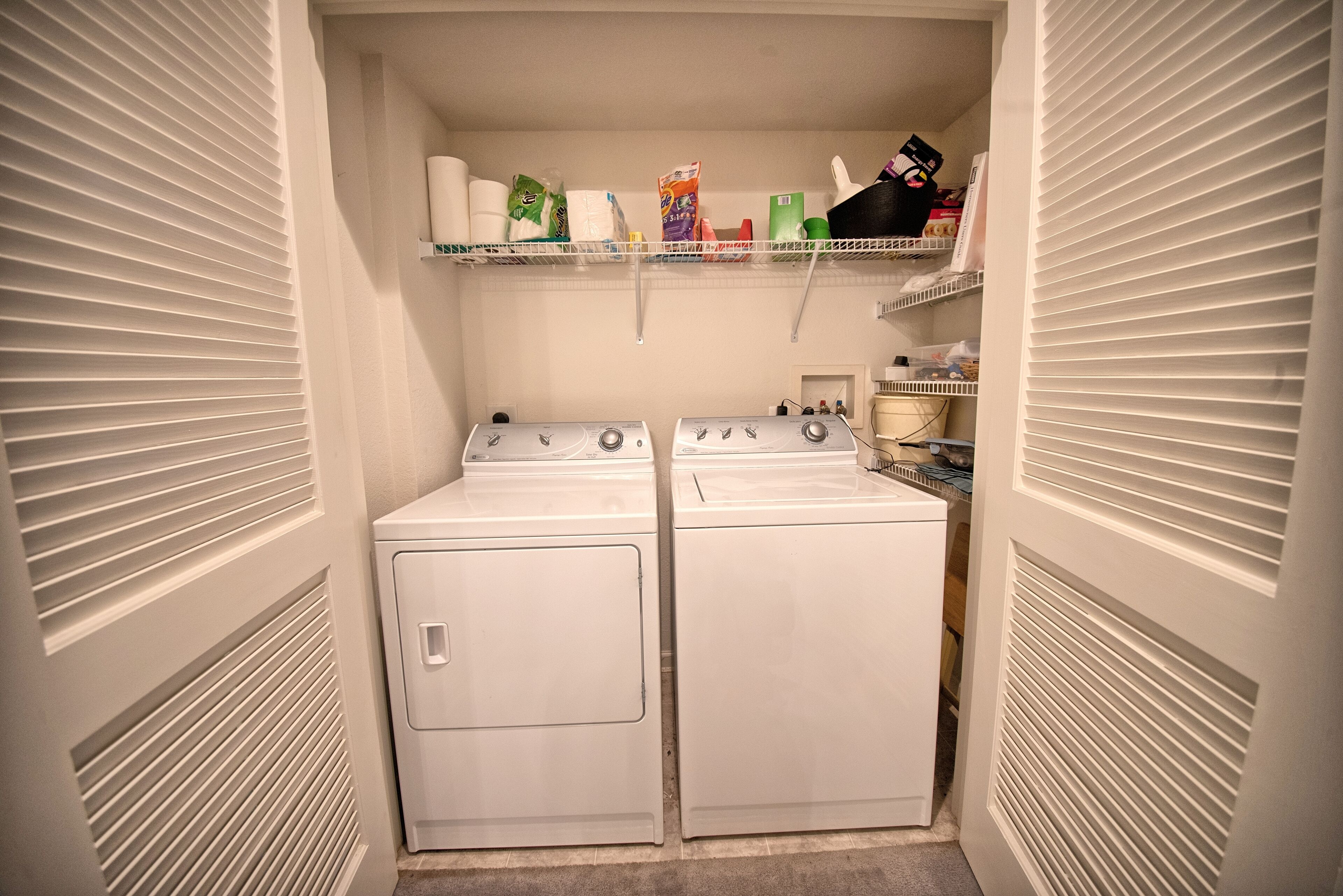 A laundry room with two white front-loading washers and dryers, shelves above holding various cleaning supplies, and a small window with blinds.