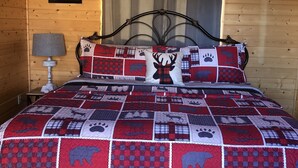Ein gemütliches Schlafzimmer mit Holzbalkendecke, einem Bett mit einer rot-weißen Steppdecke mit Tiermotiven, einer Nachttischlampe und einem Fenster mit Vorhängen.