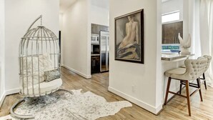 A modern living room with a hanging chair, a large painting, and a dining area with a white table and chairs. - Kirby Haus ·Modern Luxury· West U· RiceVillage · sleeps16 ·EV 🚗🔌 •3900sqft ⭐️⭐️⭐️⭐️⭐️ (Houston)