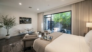 A modern hotel room with a large bed, a seating area with a sofa and chairs, a dining table, and a large window offering a view of greenery outside. - Kirby Haus ·Modern Luxury· West U· RiceVillage · sleeps16 ·EV 🚗🔌 •3900sqft ⭐️⭐️⭐️⭐️⭐️ (Houston)