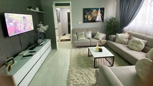 A modern living room with a flat-screen TV, a sofa, a coffee table, and a ceiling fan. - Exotic 2 Bedrooms Apartment (Accra)