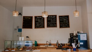 Interior de una cafetería con un menú, opciones de espresso y café helado, una máquina de café y una vitrina con pasteles.