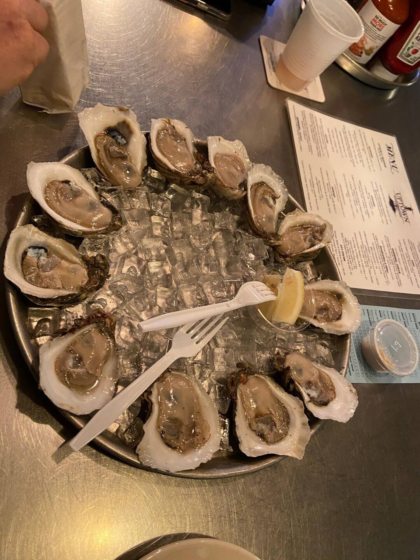 A plate of oysters on ice with a fork and lemon wedge, a menu, and condiments on a table.