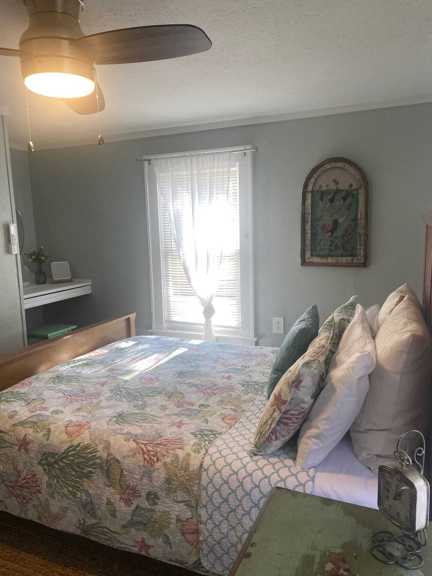 Une chambre à coucher comprenant un lit, un ventilateur de plafond, une fenêtre avec des rideaux, une étagère fixée au mur et un tableau encadré.