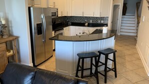 Cocina moderna con armarios blancos, refrigerador de acero inoxidable, encimera negra y una pequeña isla con taburetes.