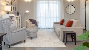 Un salón con un sillón a cuadros de galgo, un sofá con cojines naranjas, una mesita y un amplio ventanal con cortinas de encaje.