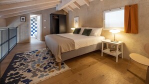Ein Schlafzimmer mit Bett, Nachttischen, einem Fenster mit Vorhängen, einer Holzbalkendecke und Blick ins Badezimmer.