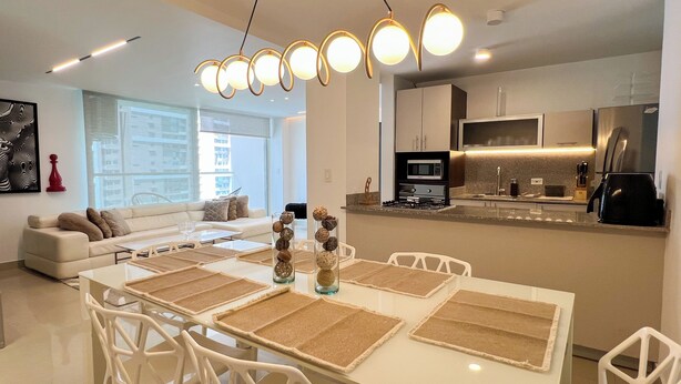 A modern kitchen with a central island, a dining area with a white table and chairs, and a living room with a sofa and a large window. - Accommodation with views in Costa del Este 6 (Panama)